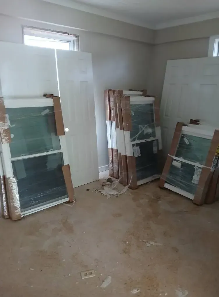 Stacked window panes and doors leaning against walls in a bare room with a dusty floor