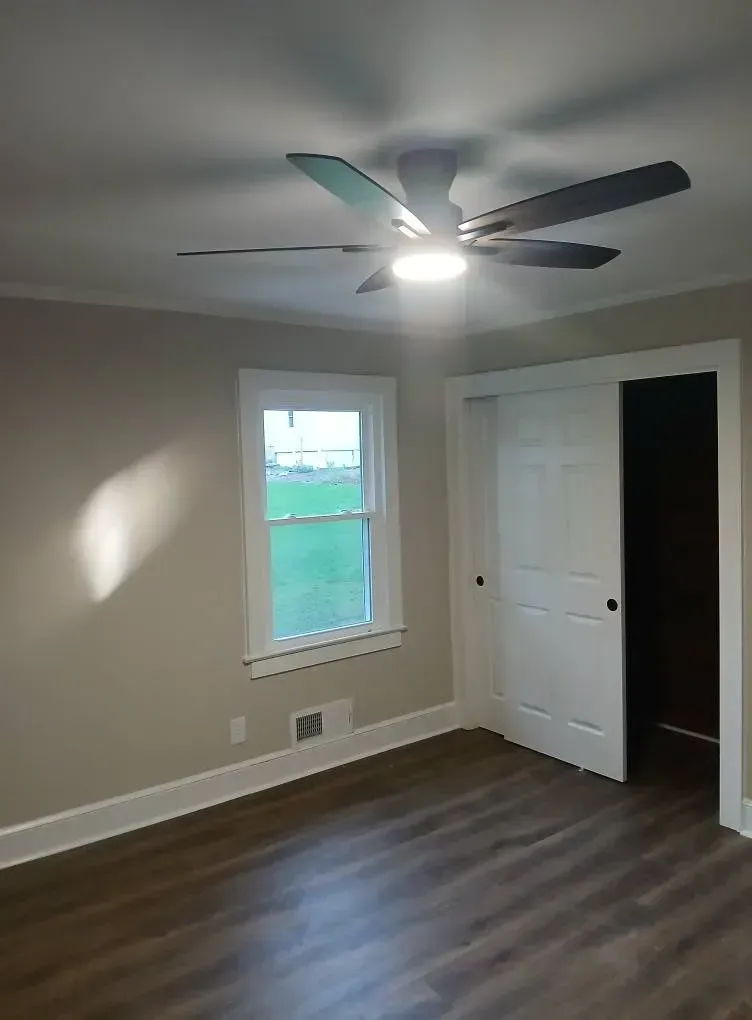 Empty beige bedroom with ceiling fan, window, closet doors, and wood-look flooring.