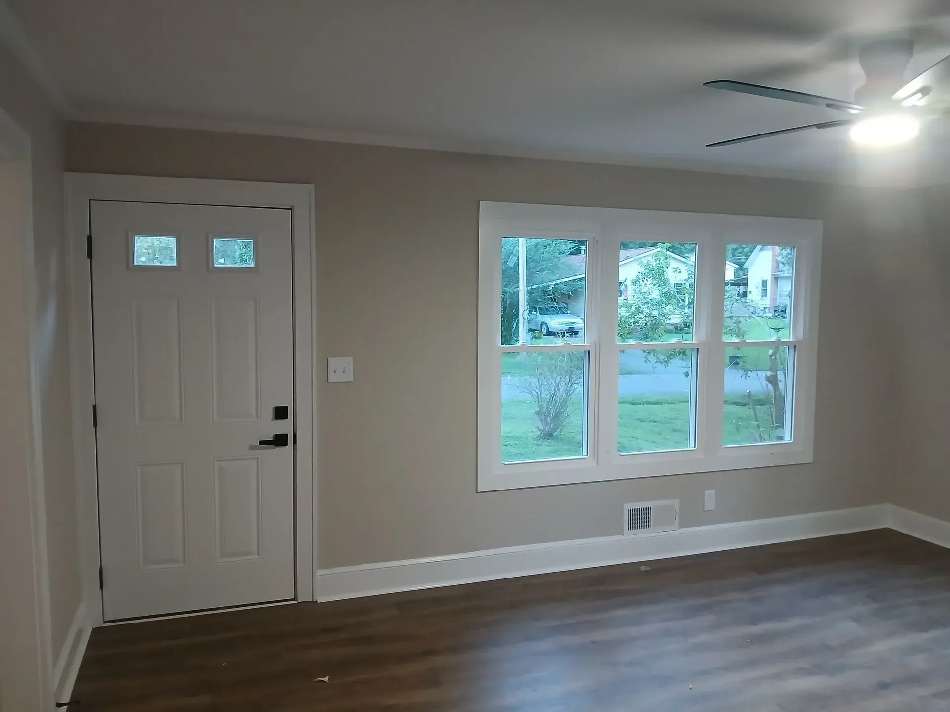 Empty living room with beige walls, white front door, large window, and dark wood floors.
