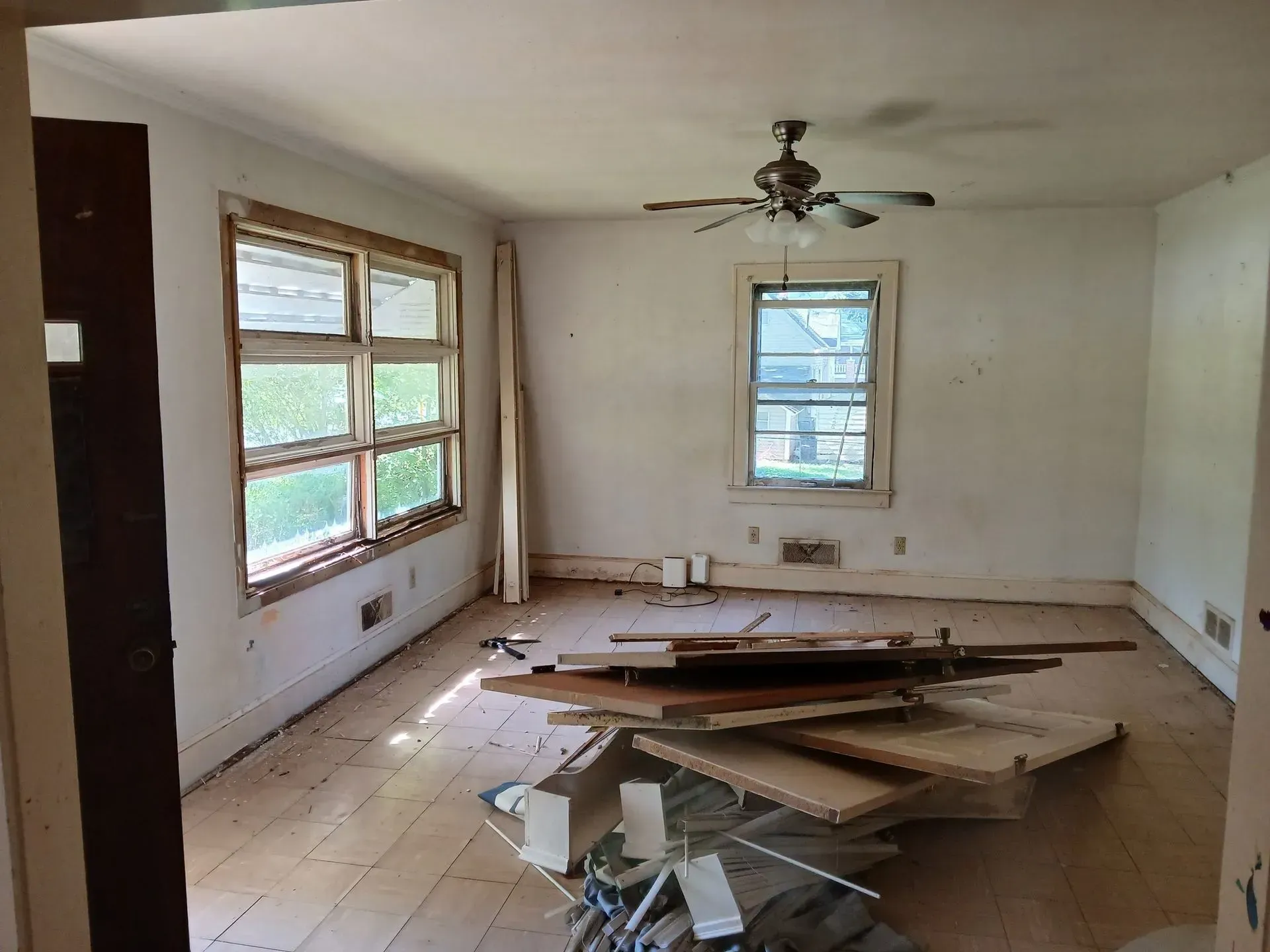 Abandoned room with debris on the floor, bare walls, windows, and a ceiling fan.