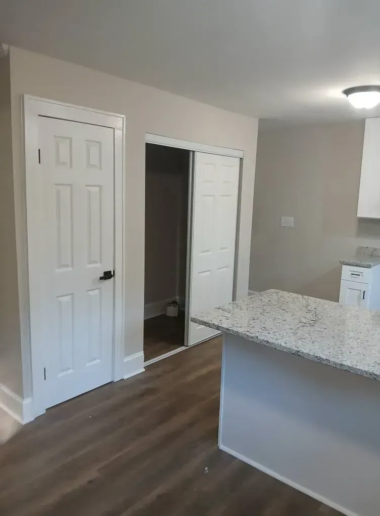 Empty kitchen with granite island, white cabinets, wood floors, and a dark doorway in the background
