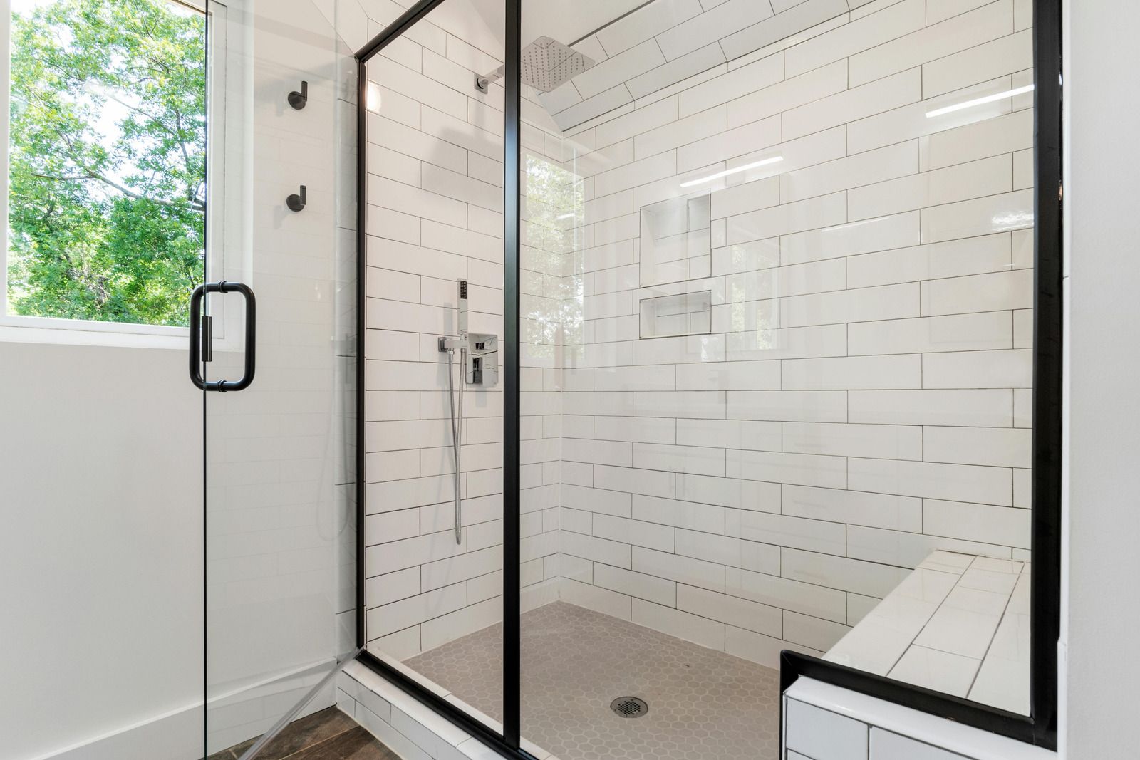 Modern white-tiled shower with glass door and black hardware, beside a window and bench seat