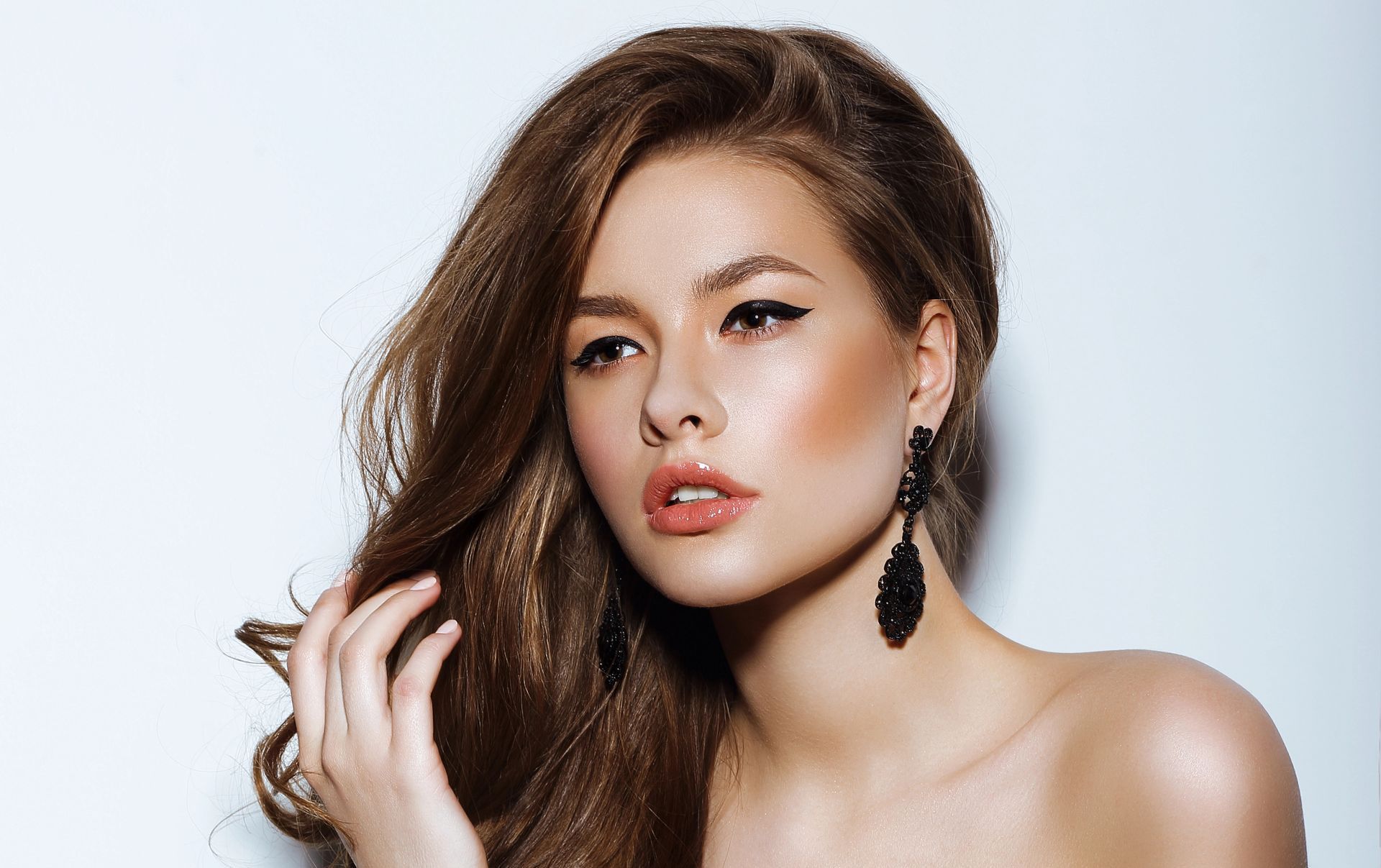 A woman with long brown hair and black earrings is standing in front of a white background.