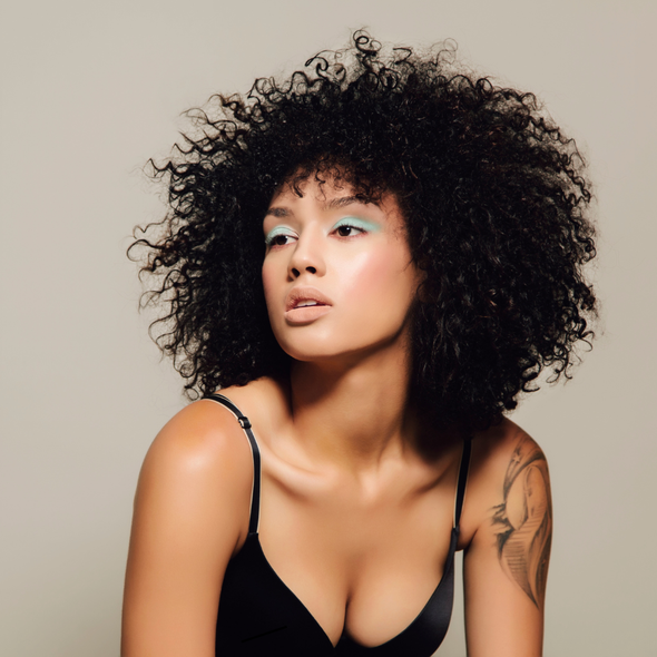 A model with curly hair wearing a black bra sitting down.