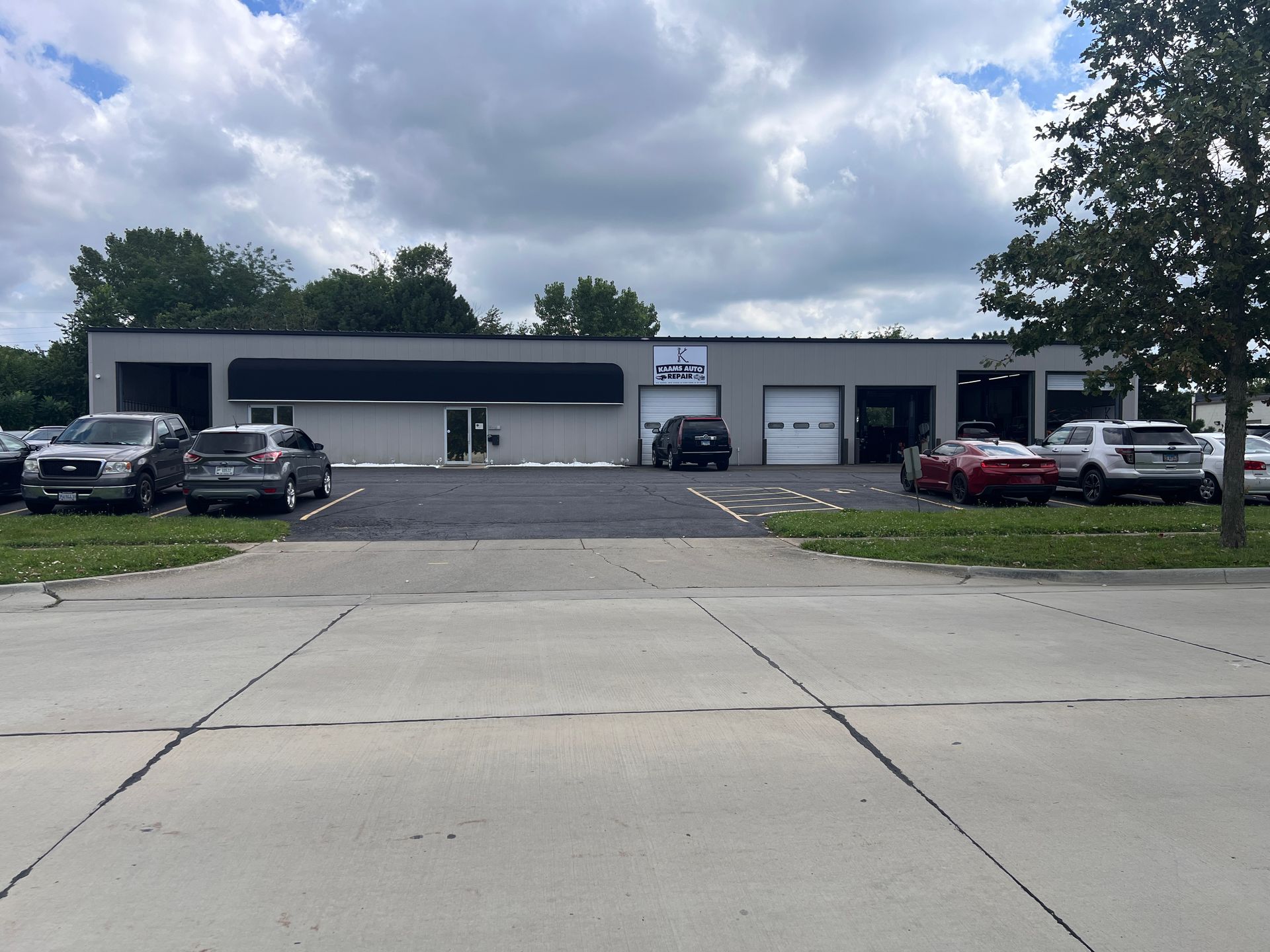 Cars parked outside a gray commercial building with a black awning and two garage doors under a cloudy sky. | KAAMS Auto Repair