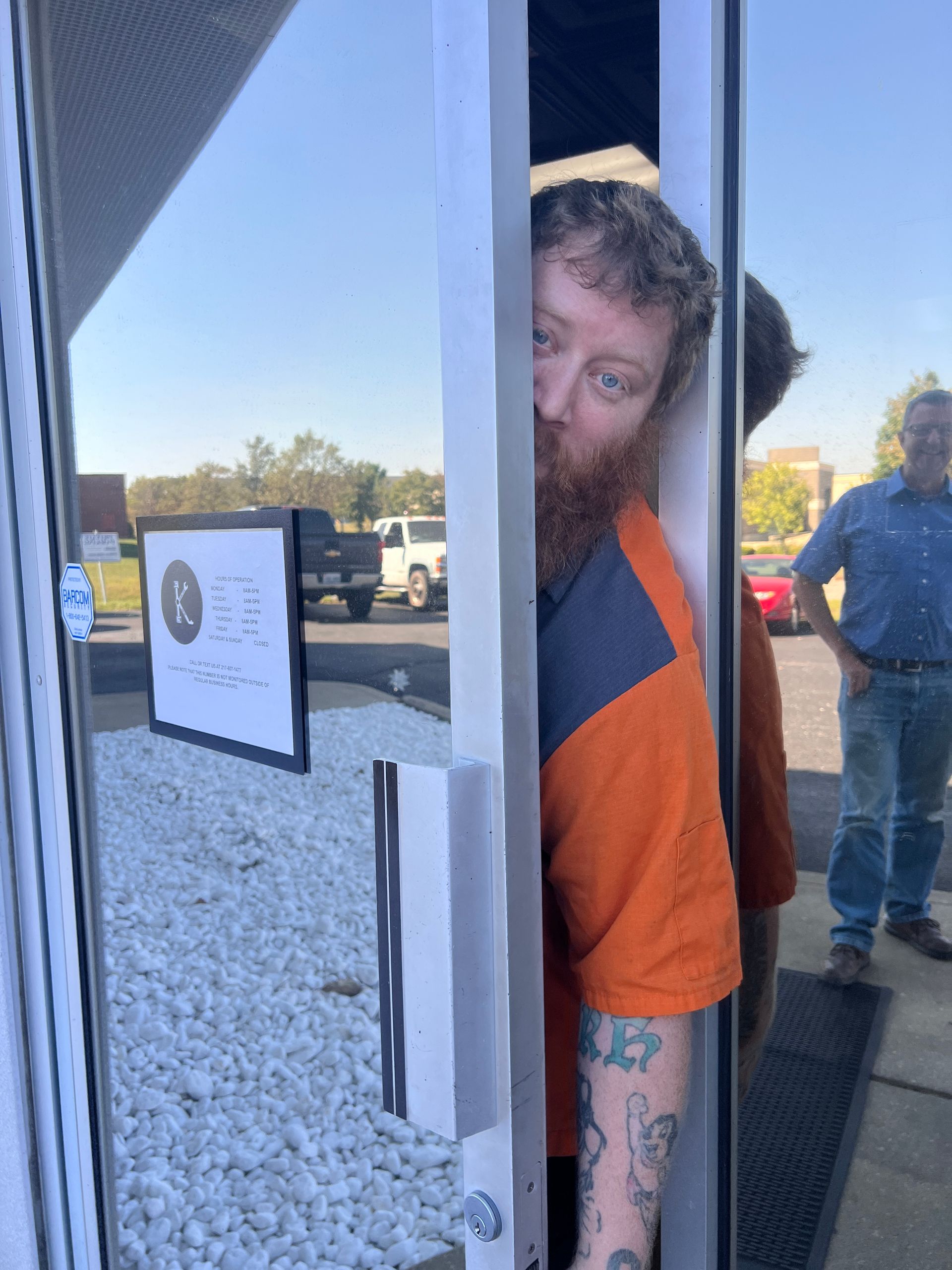 Man peeking around a glass door. He has a red beard, orange shirt, and arm tattoos. | KAAMS Auto Repair