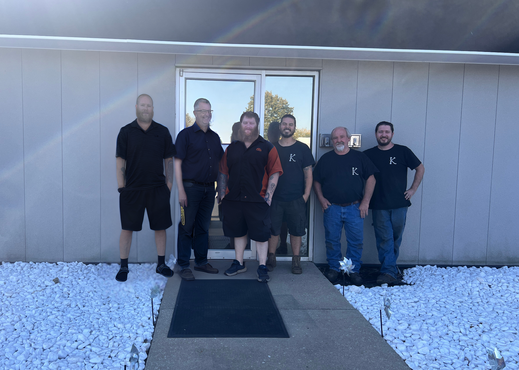 Six men stand outside a building with glass doors. They are smiling. White rocks line the walkway. | KAAMS Auto Repair
