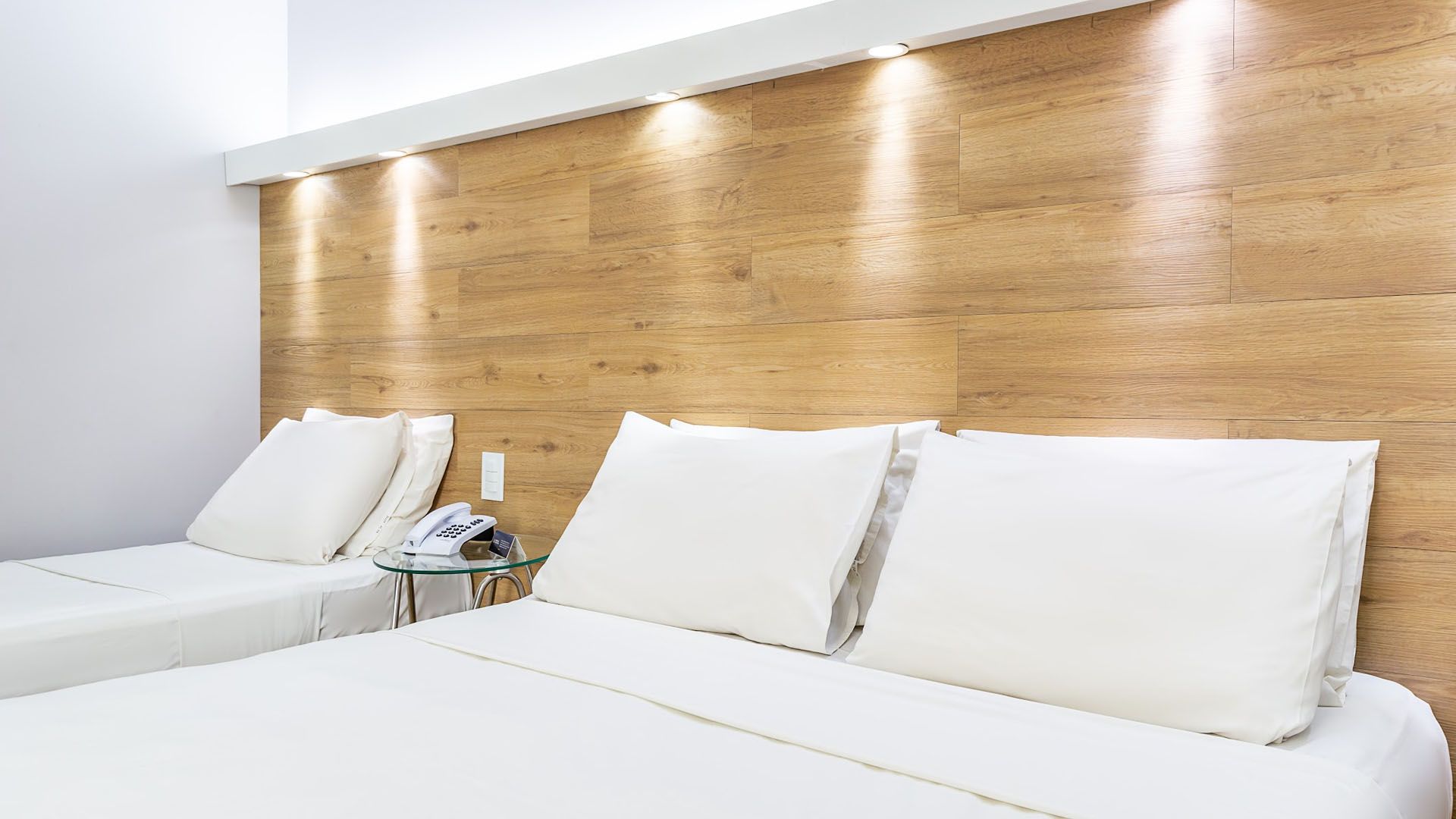 Quarto com três camas brancas e cabeceira de madeira com iluminação embutida.