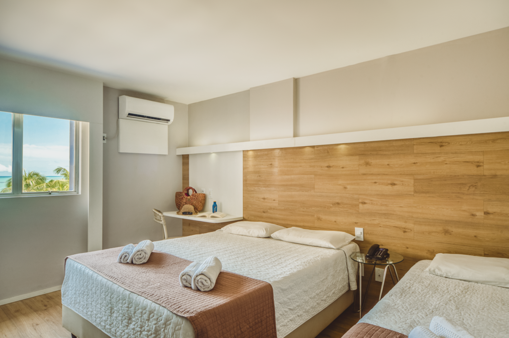 Quarto de hotel com duas camas, janela com vista para o mar, cabeceira de madeira clara e uma pequena escrivaninha.