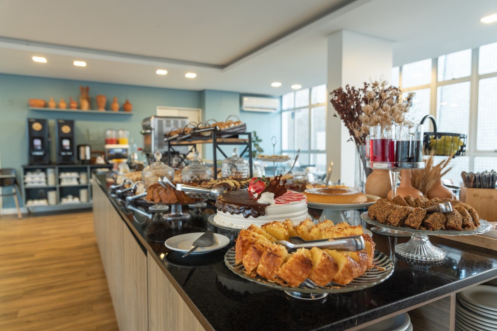 Buffet com diversos bolos, doces e bebidas expostos em um ambiente moderno e iluminado.