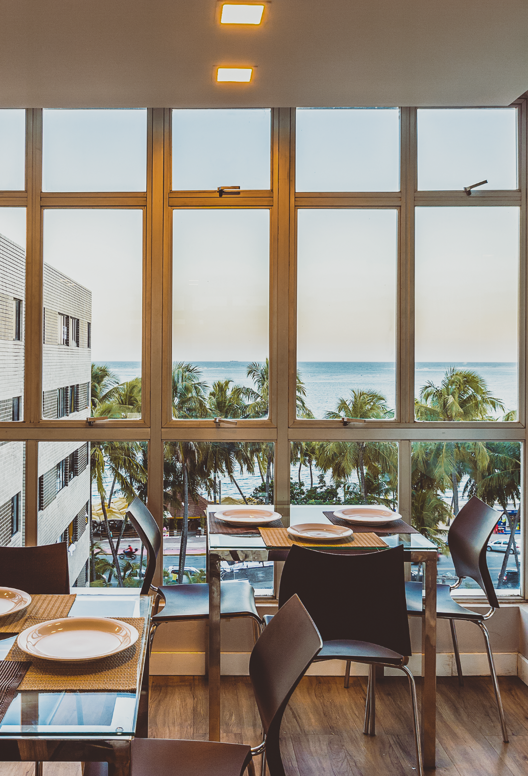 Mesa de jantar posta para quatro pessoas perto de uma grande janela com vista para o oceano e as palmeiras.