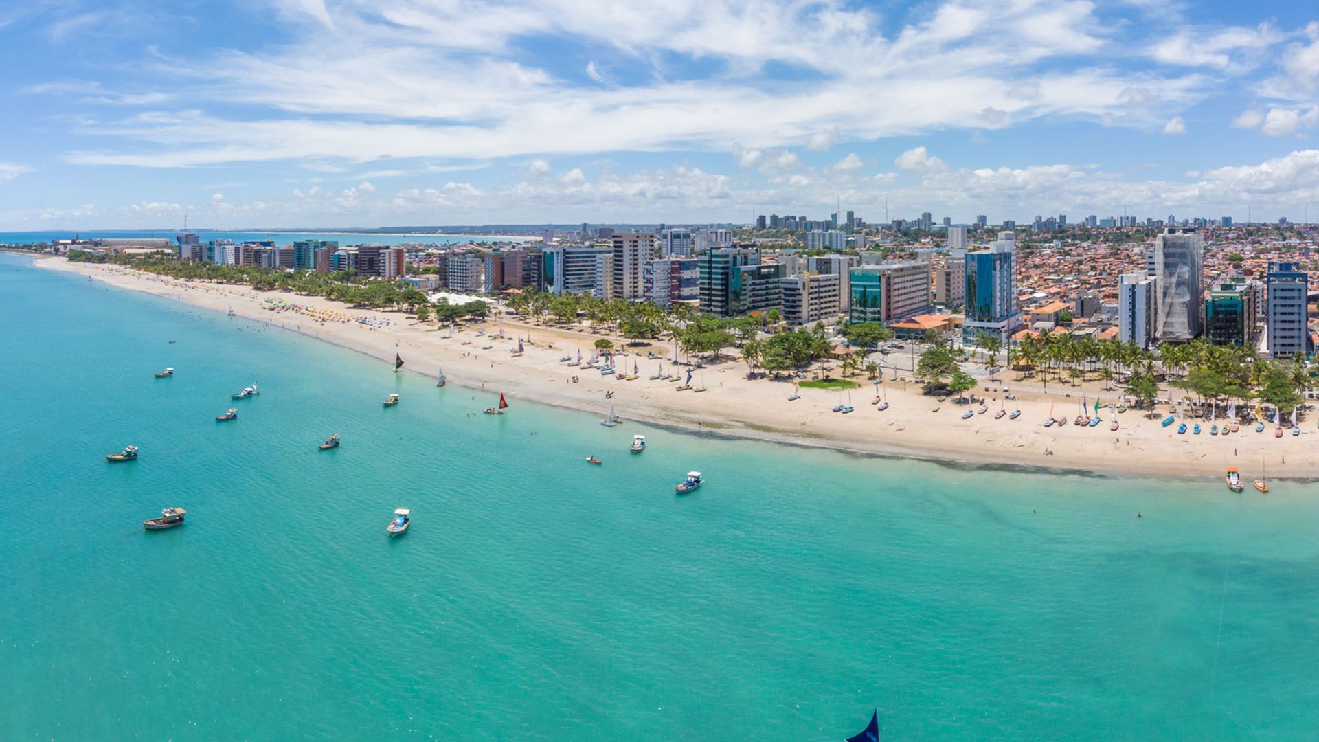 Vista aérea de uma praia ensolarada em Natal, Brasil, com águas azul-turquesa, barcos e o horizonte da cidade.
