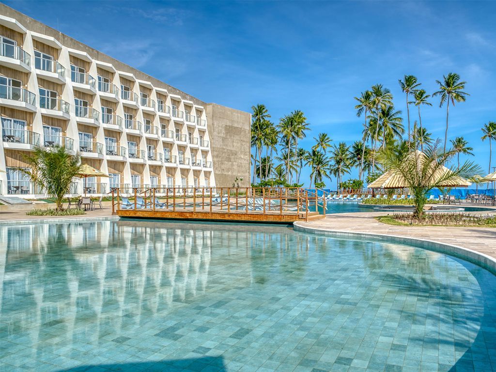 Hotel com piscina, ponte e palmeiras sob um céu azul.