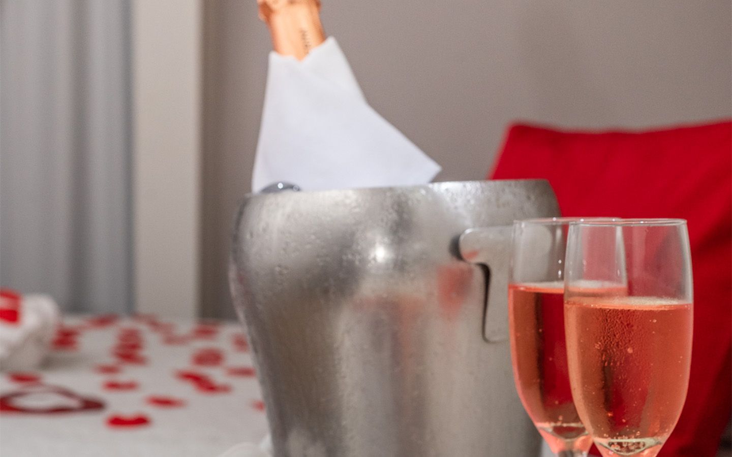 Balde de champanhe com duas taças de champanhe rosé; cama com pétalas de rosas vermelhas.