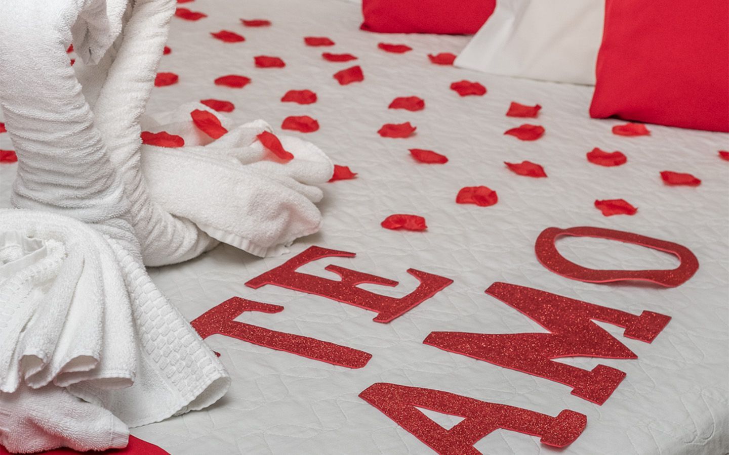 Champanhe em balde de gelo com duas taças cheias, cama vermelha e pétalas de rosa.