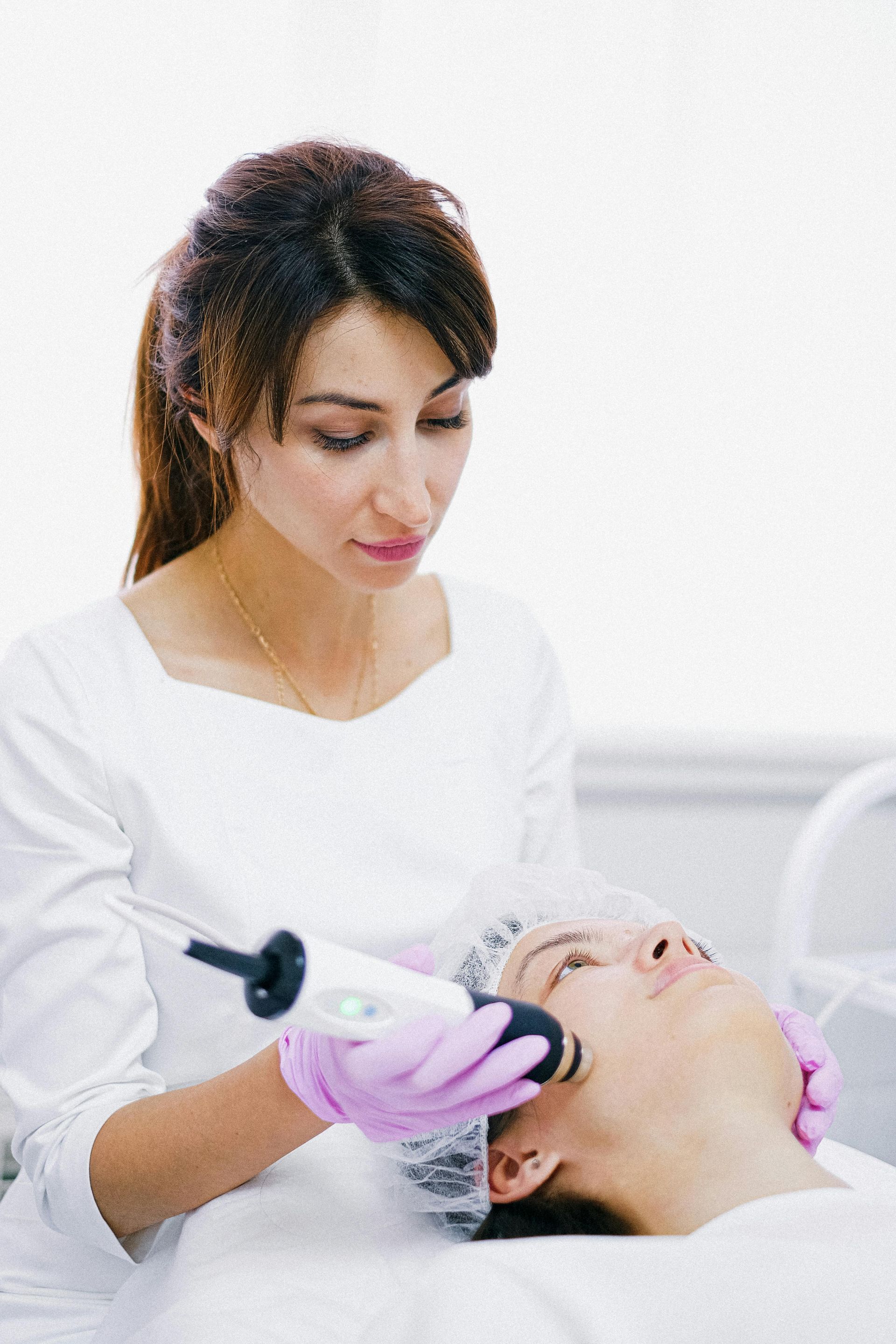 A beautician with pink gloves performs a facial treatment on a client. The client lies with a hairnet. The setting appears to be a clinic.
