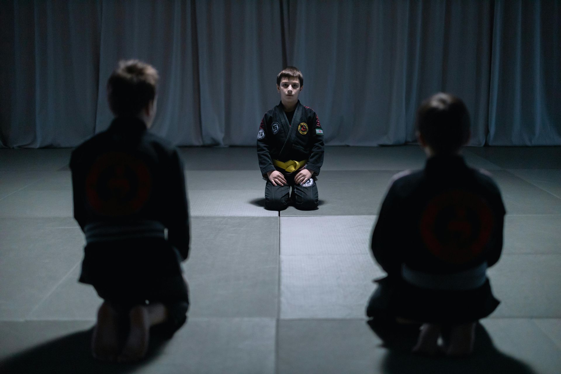 Three young martial artists kneeling, facing center, in a training room.