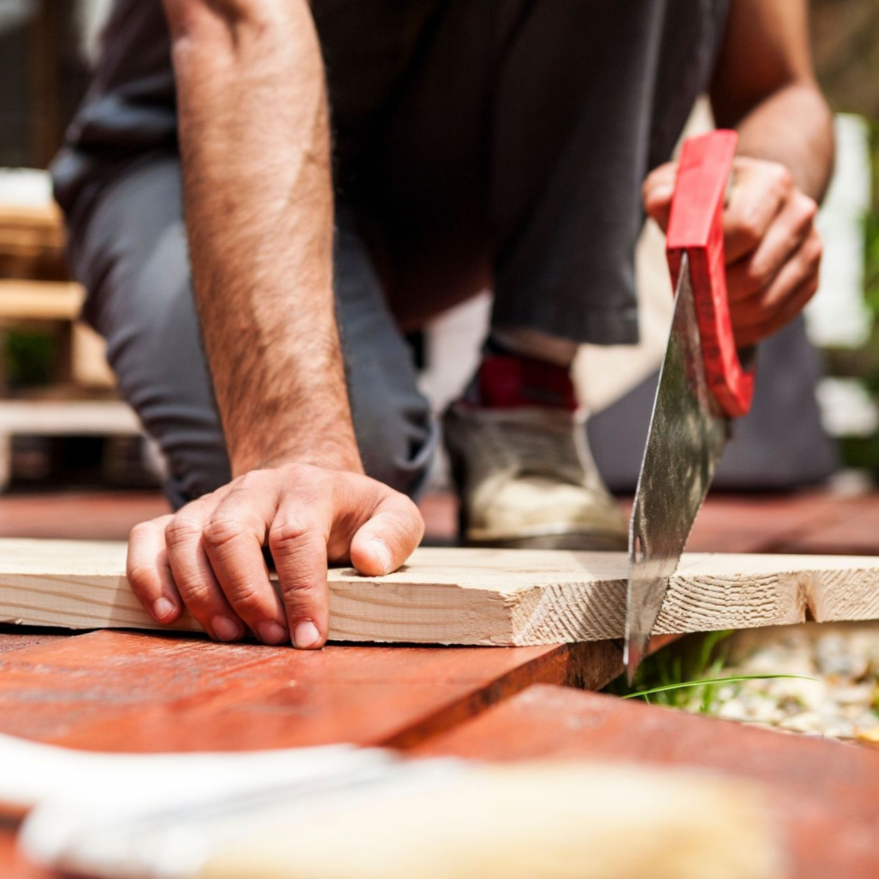 Metal Roof Person sawing a wooden plank with a red-handled hand saw on a red surface.