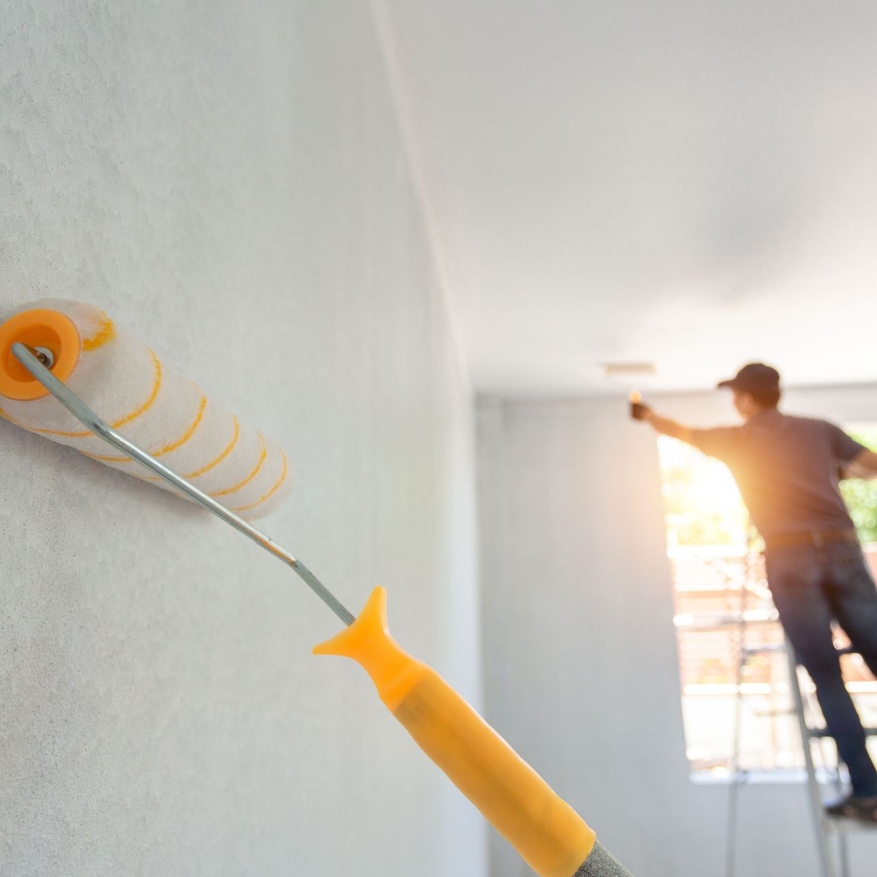 Metal Roof A person paints a wall and ceiling with a roller, standing on a ladder in a room.