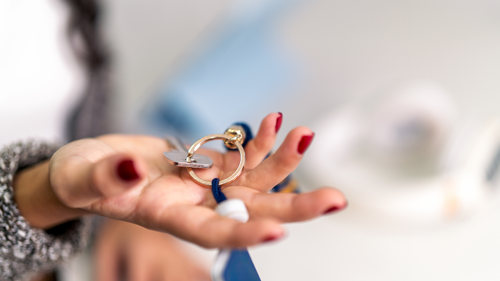 Hand holding a gold keyring with a blue lanyard, red nail polish.