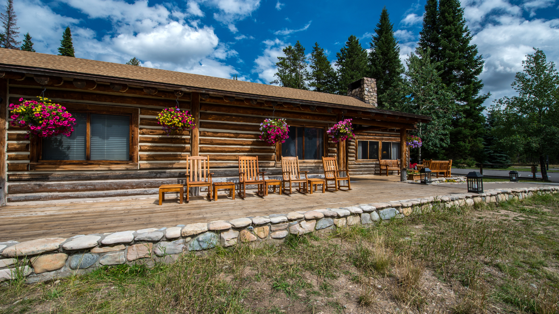 Log cabin with hanging flower baskets and wooden chairs on a porch, blue sky and trees in the background.