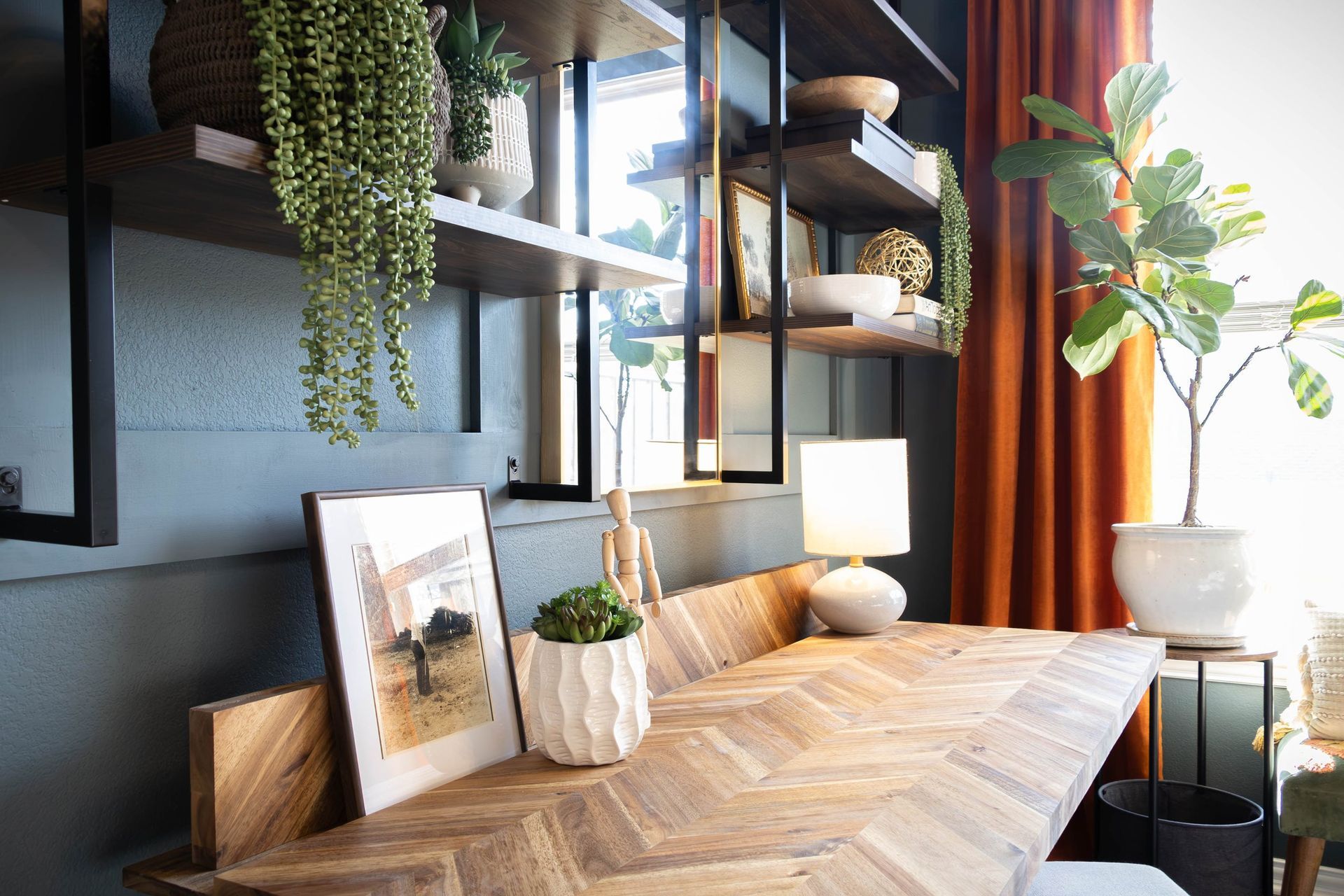 A room with a wooden table , shelves , plants and a lamp.