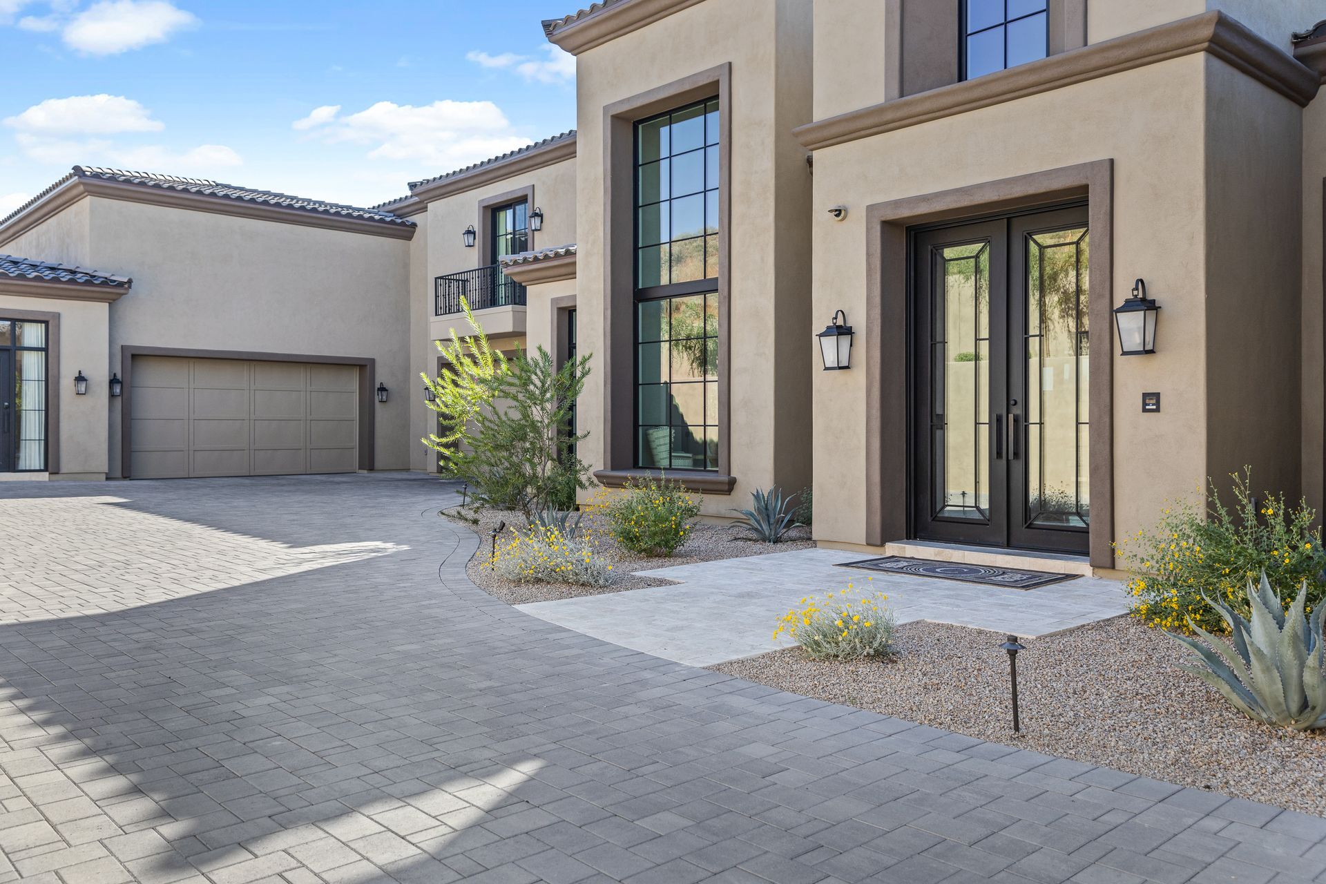 Driveway and Entrance: A modern two-story home with a beige exterior and large, black-framed windows. The entrance features double glass doors with black frames, flanked by two outdoor wall lanterns. The driveway is paved with gray bricks, leading up to a double garage door. The front yard has desert landscaping with small shrubs, cacti, and flowering plants. The sky is clear and blue, adding to the bright and welcoming appearance of the property.