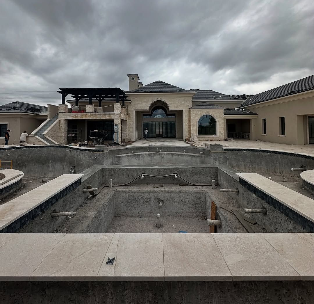 Luxury construction site in Arizona showing a large, unfinished swimming pool in the foreground of a luxury home under a cloudy sky.