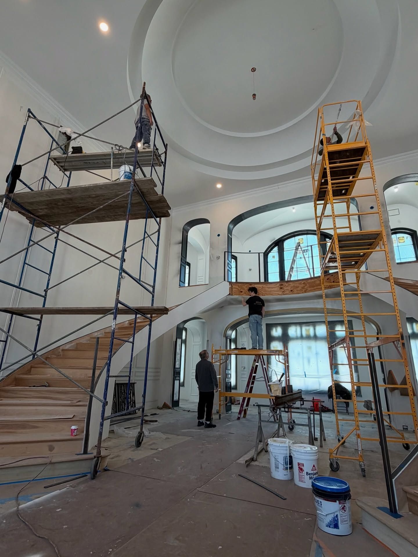 Construction workers on scaffolding inside a high-ceilinged room with a dual curved staircase and double height ceilings