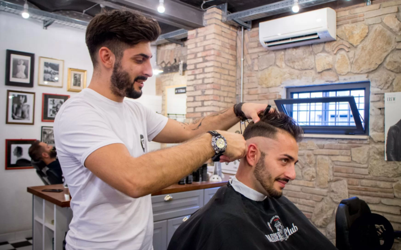 Un barbiere taglia i capelli a un cliente in una moderna barberia con pareti in mattoni e foto incorniciate.