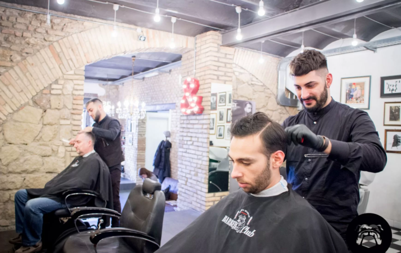 Barbiere con due clienti che si fanno tagliare i capelli sotto luci intense, con muri in pietra a vista sullo sfondo.