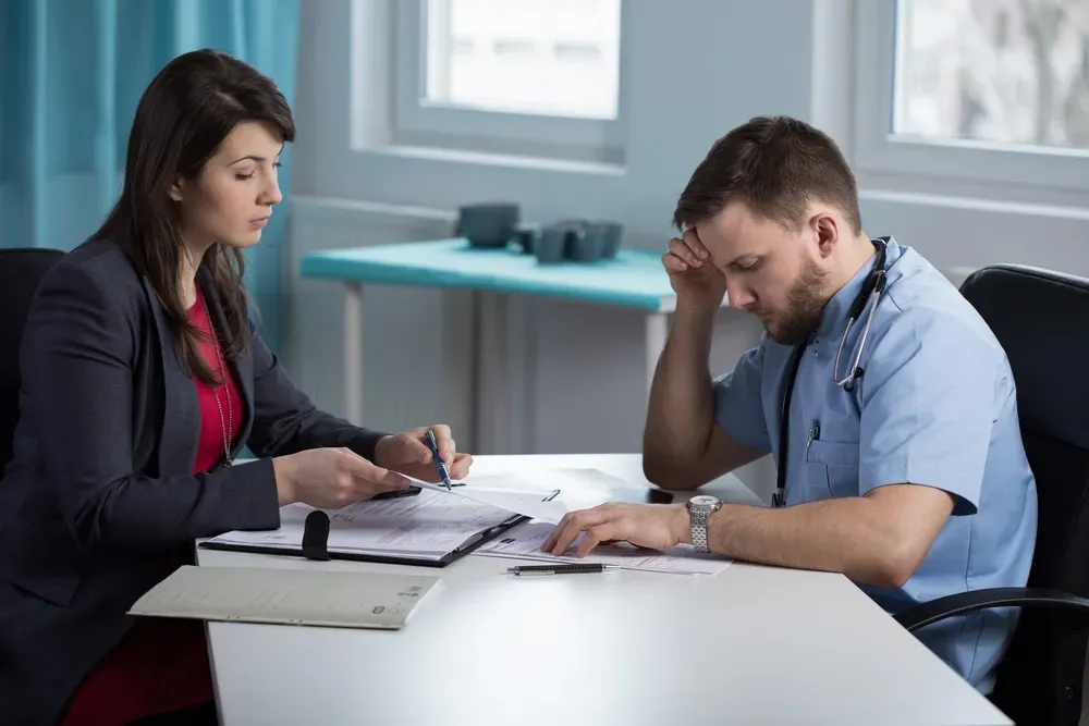 Woman in suit showing documents to a doctor, who looks stressed. They sit at a desk in a medical office. — Dwyer Law Group in Surfers Paradise, QLD