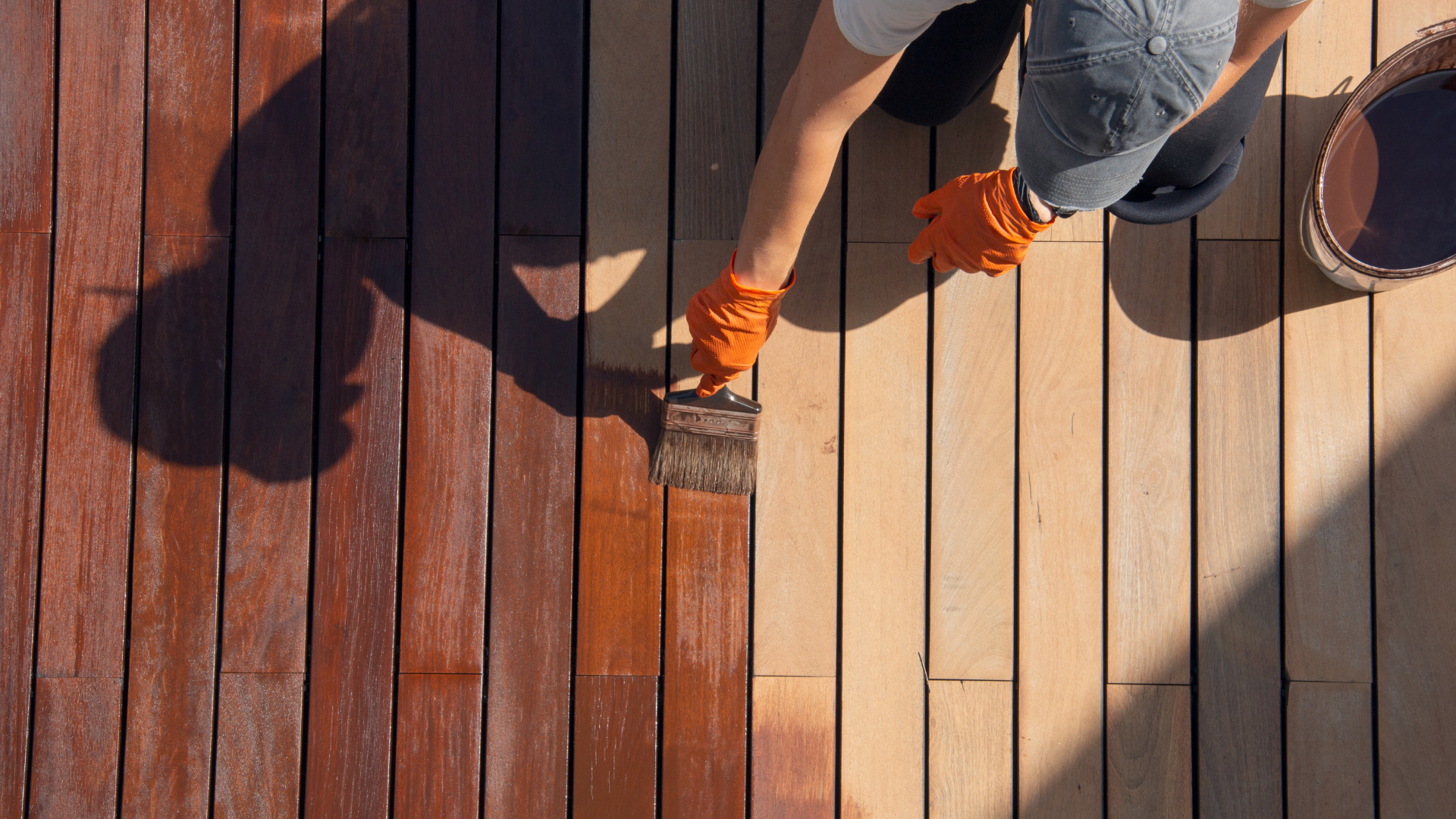 Person painting a wooden deck, with fresh dark stain beside unfinished tan boards.