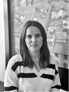 Frau mit schulterlangem Haar in einem gestreiften Pullover vor einem verschwommenen Stadtbildgemälde.