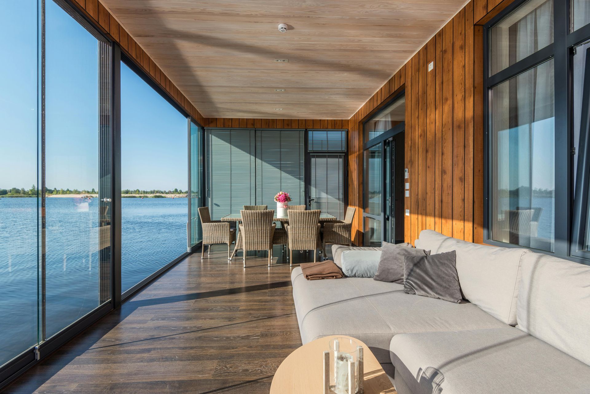 Wooden-paneled room with floor-to-ceiling windows overlooking water, furnished with a sofa and outdoor dining set.