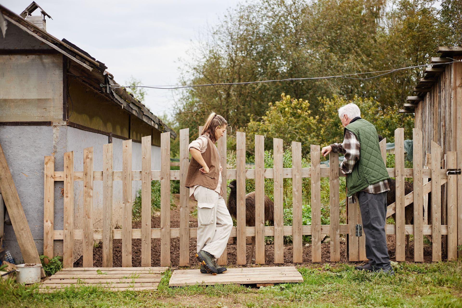 Woman and older man standing near a wooden fence, talking outdoors. Overcast sky, greenery in background.