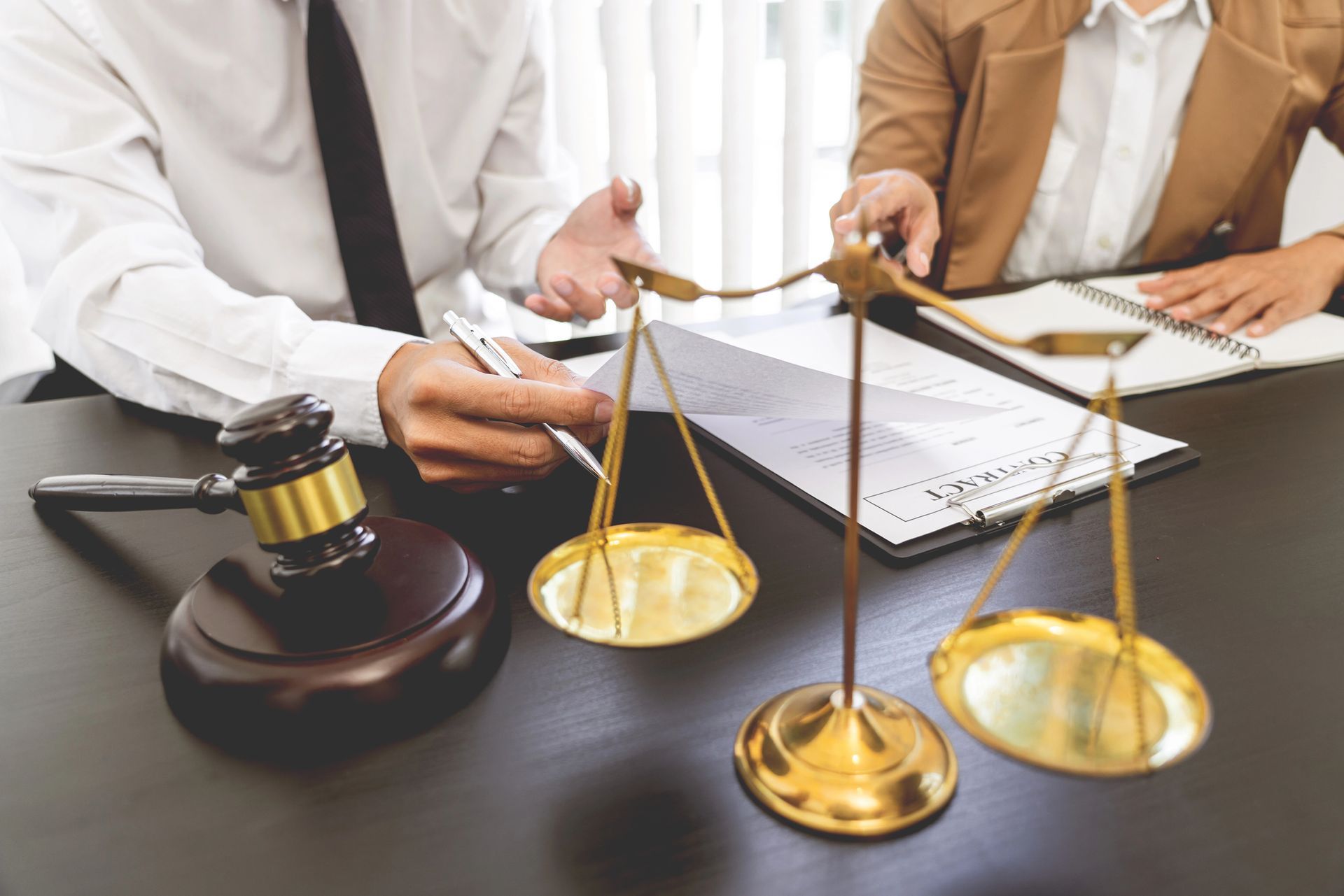 Due persone sedute a una scrivania, con una bilancia e un martelletto. Una indica un documento.