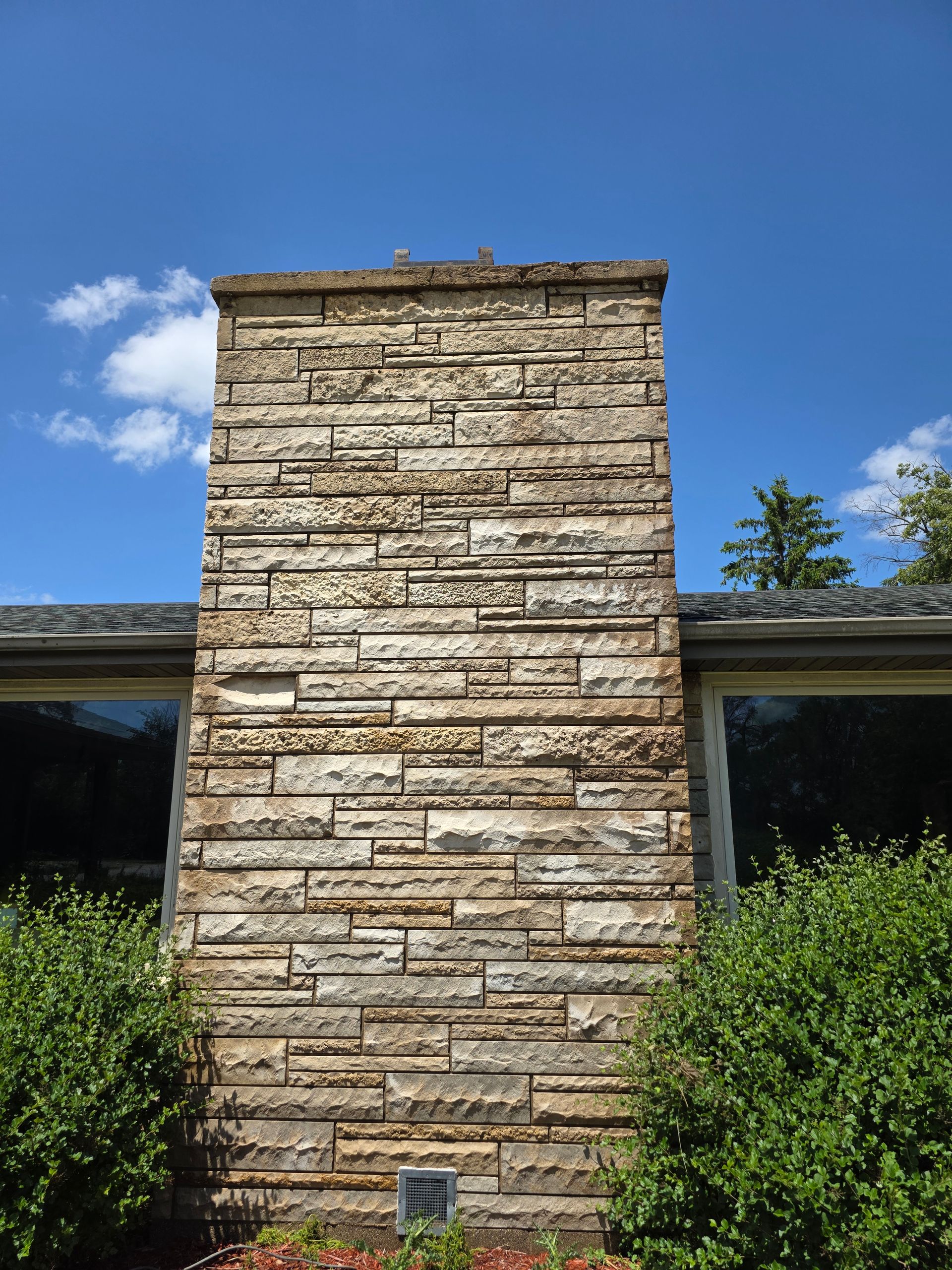 Stone chimney against blue sky, flanked by bushes and windows.