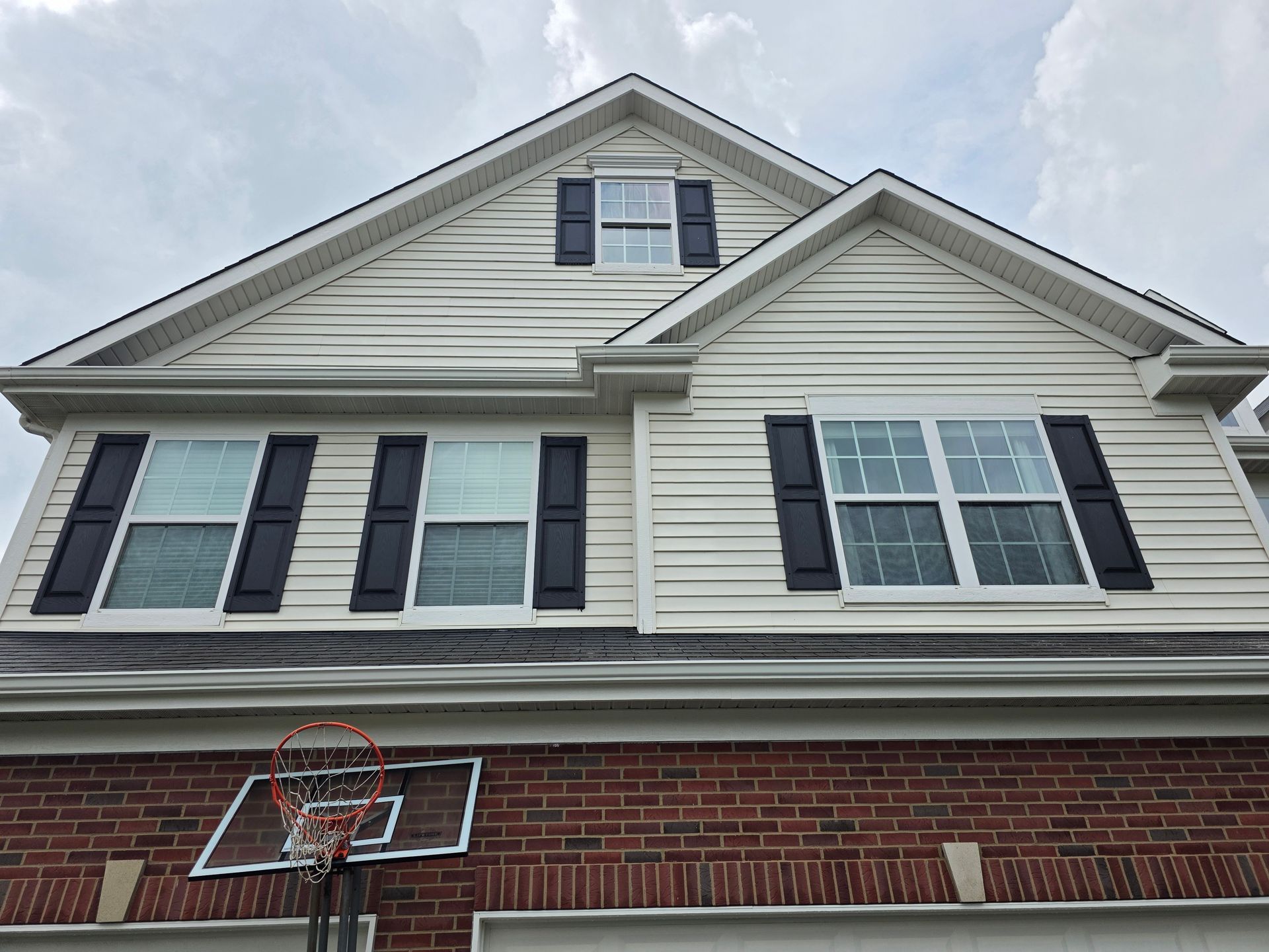 Two-story house with white siding and black shutters on windows, brick base, and a basketball hoop attached.