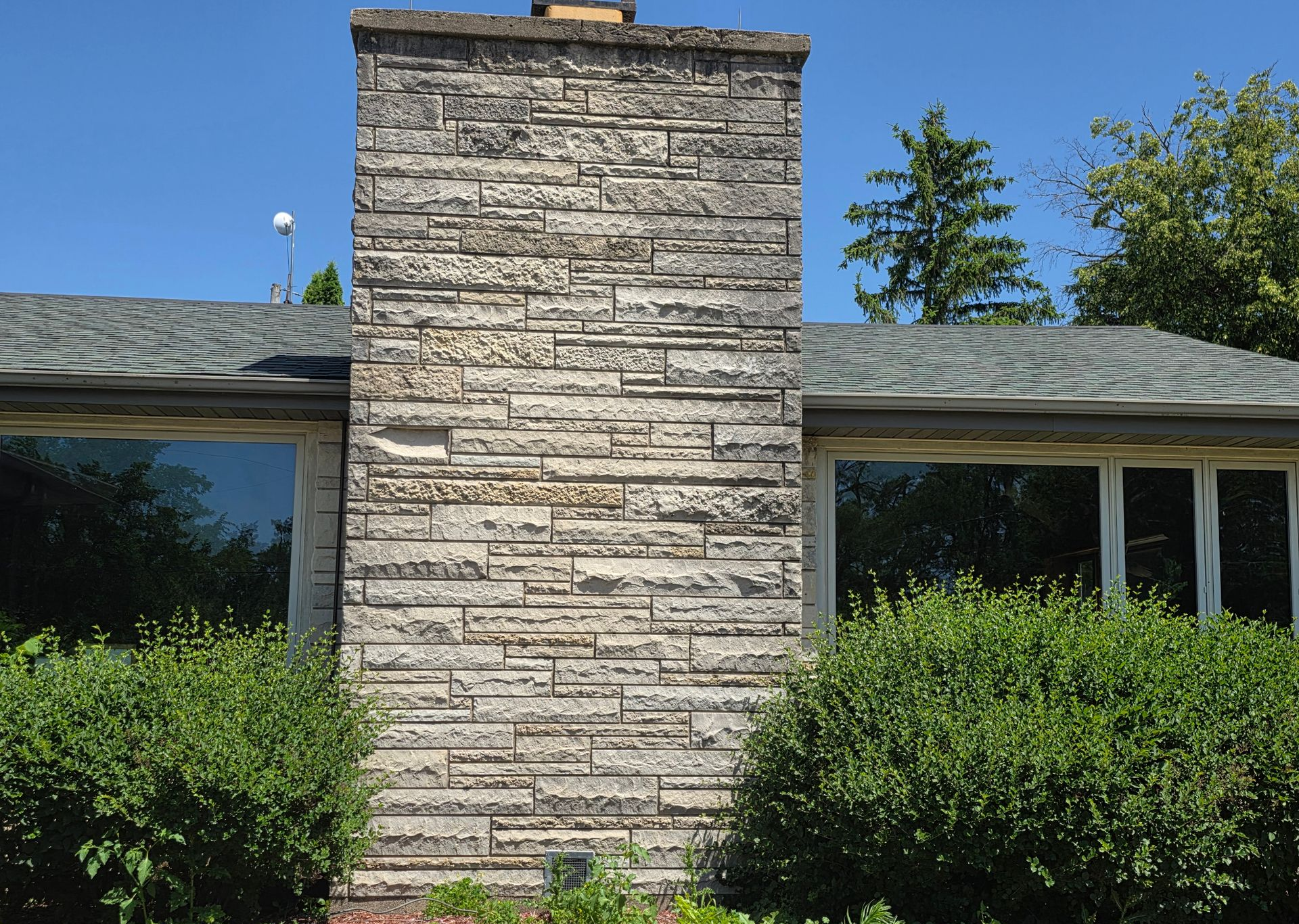 Stone chimney on a house with large windows and green bushes in front.