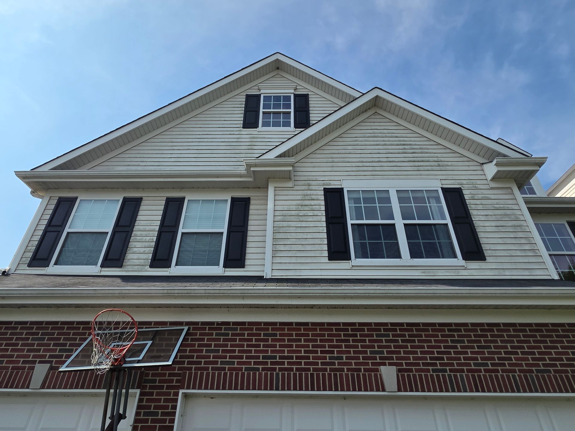 Two-story house with white siding, black shutters, brick base, basketball hoop. Sunny, blue sky background.