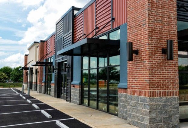 Row of retail storefronts with brick and metal facades and awnings, in a parking lot.