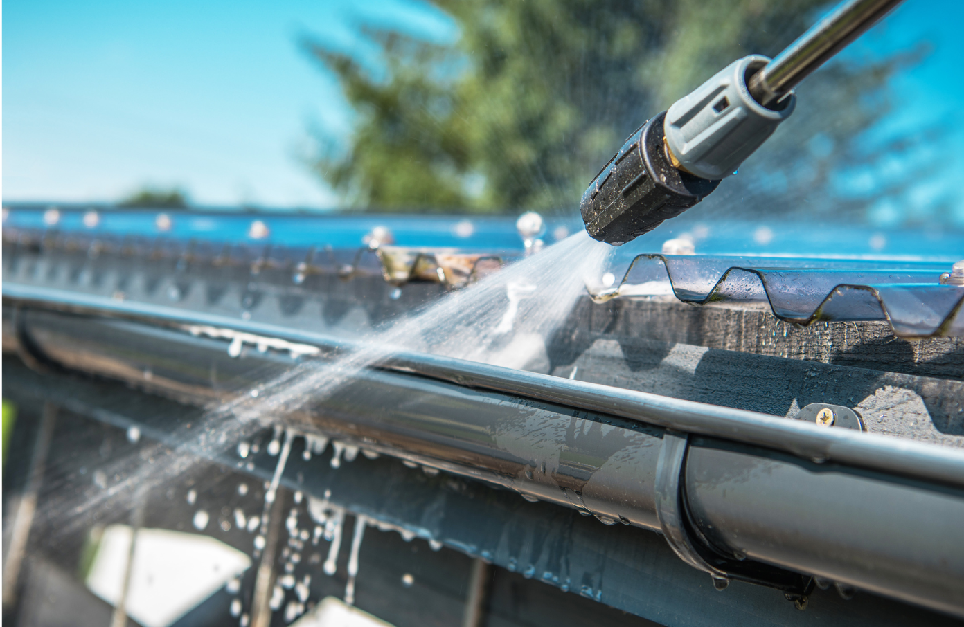 A pressure washer cleaning a black gutter filled with leaves. Blue sky in background.