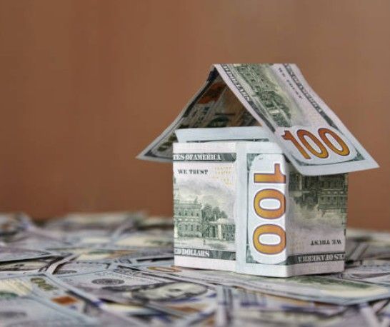 House made of US hundred-dollar bills on a pile of cash, against a brown background.