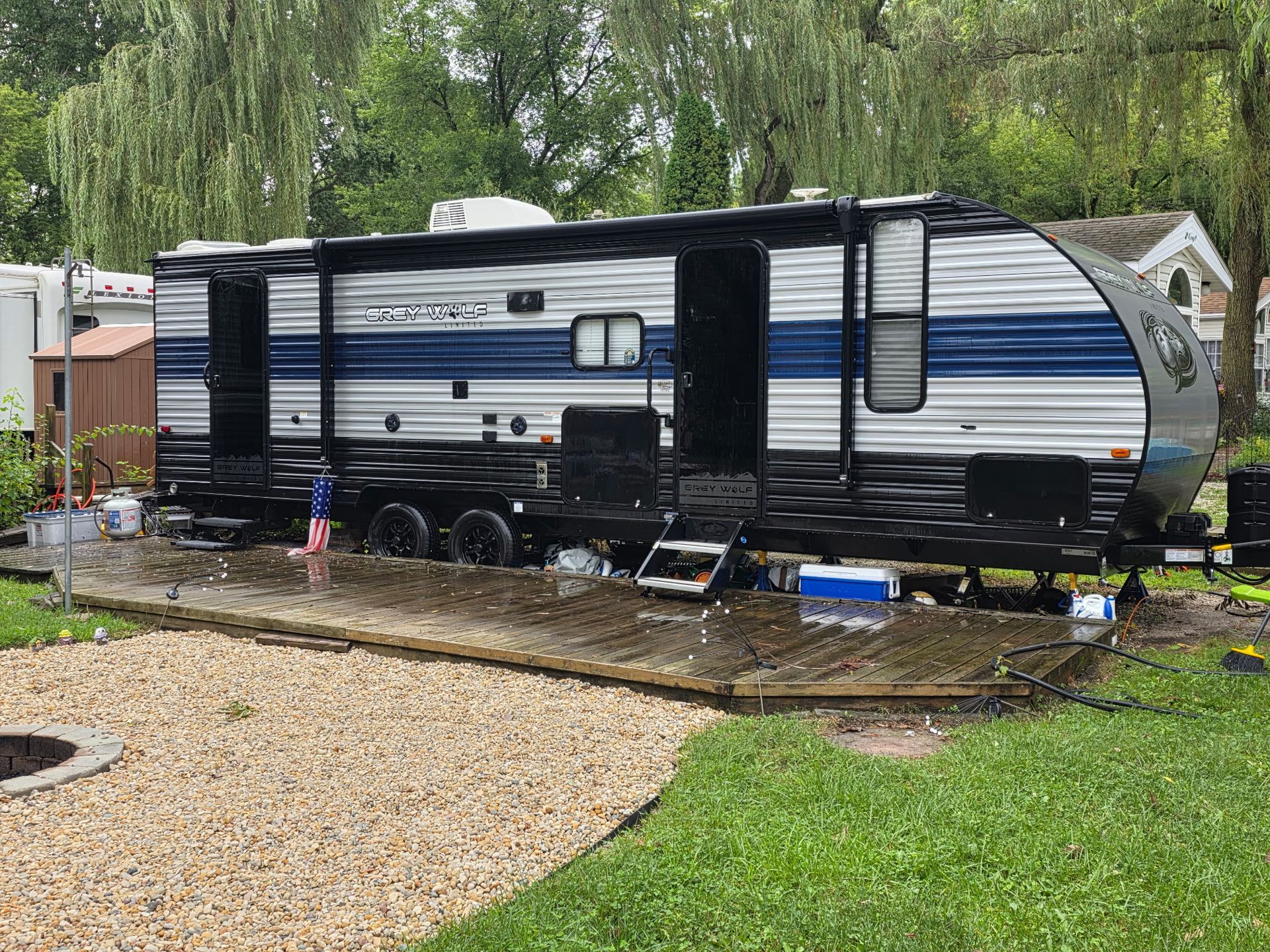 RV parked on a wooden deck with rain. Exterior is blue and black. Gravel and grass in the foreground.
