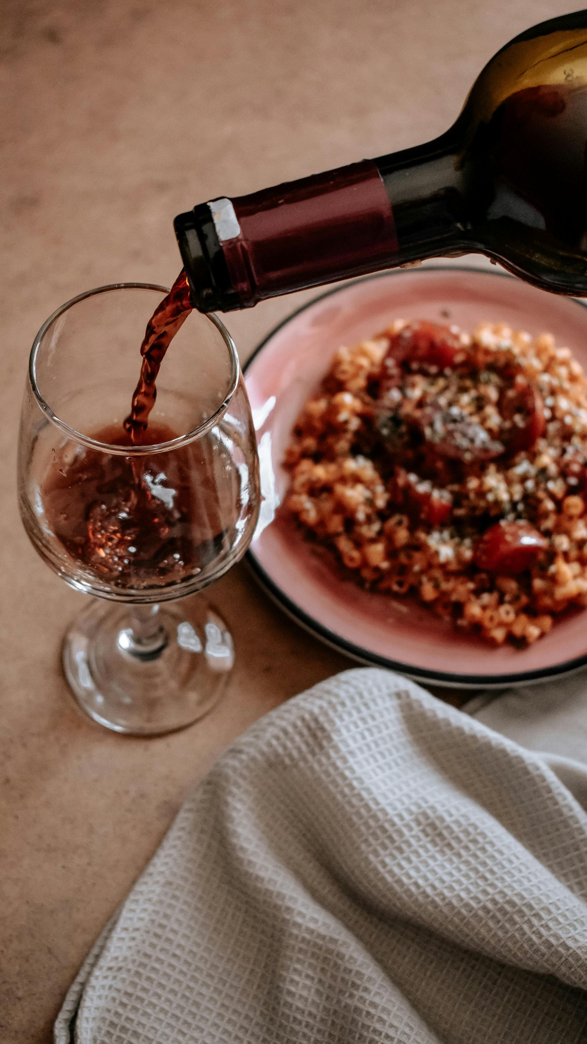 Wine being poured into a glass beside a plate of food. Gray cloth on the side, neutral background.