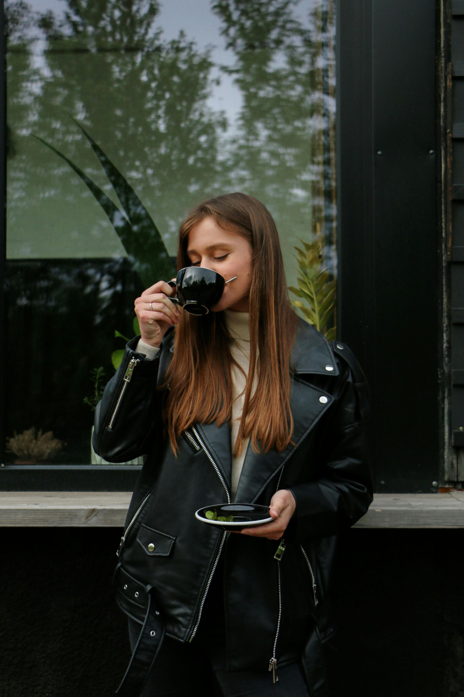 Woman in leather jacket drinks from black cup outdoors by window.