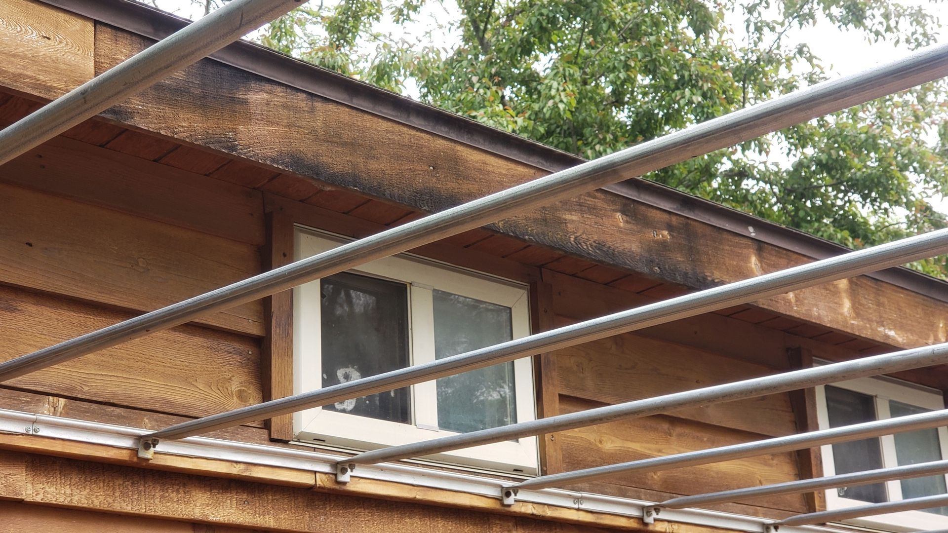 Brown wooden building with a white-framed window, metal scaffolding in the foreground, and trees in the background.