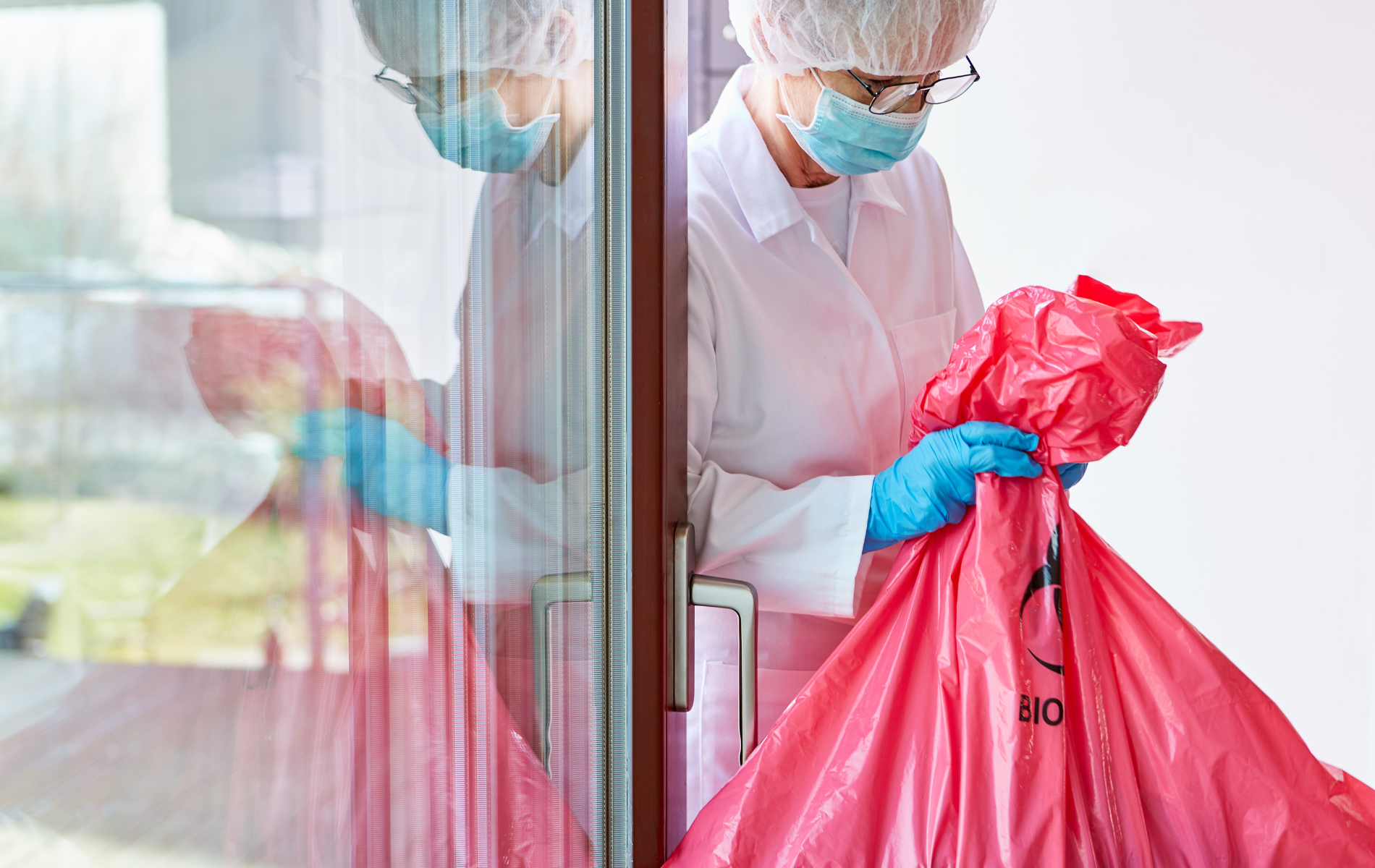 A woman in a lab coat and mask is holding a red trash bag.