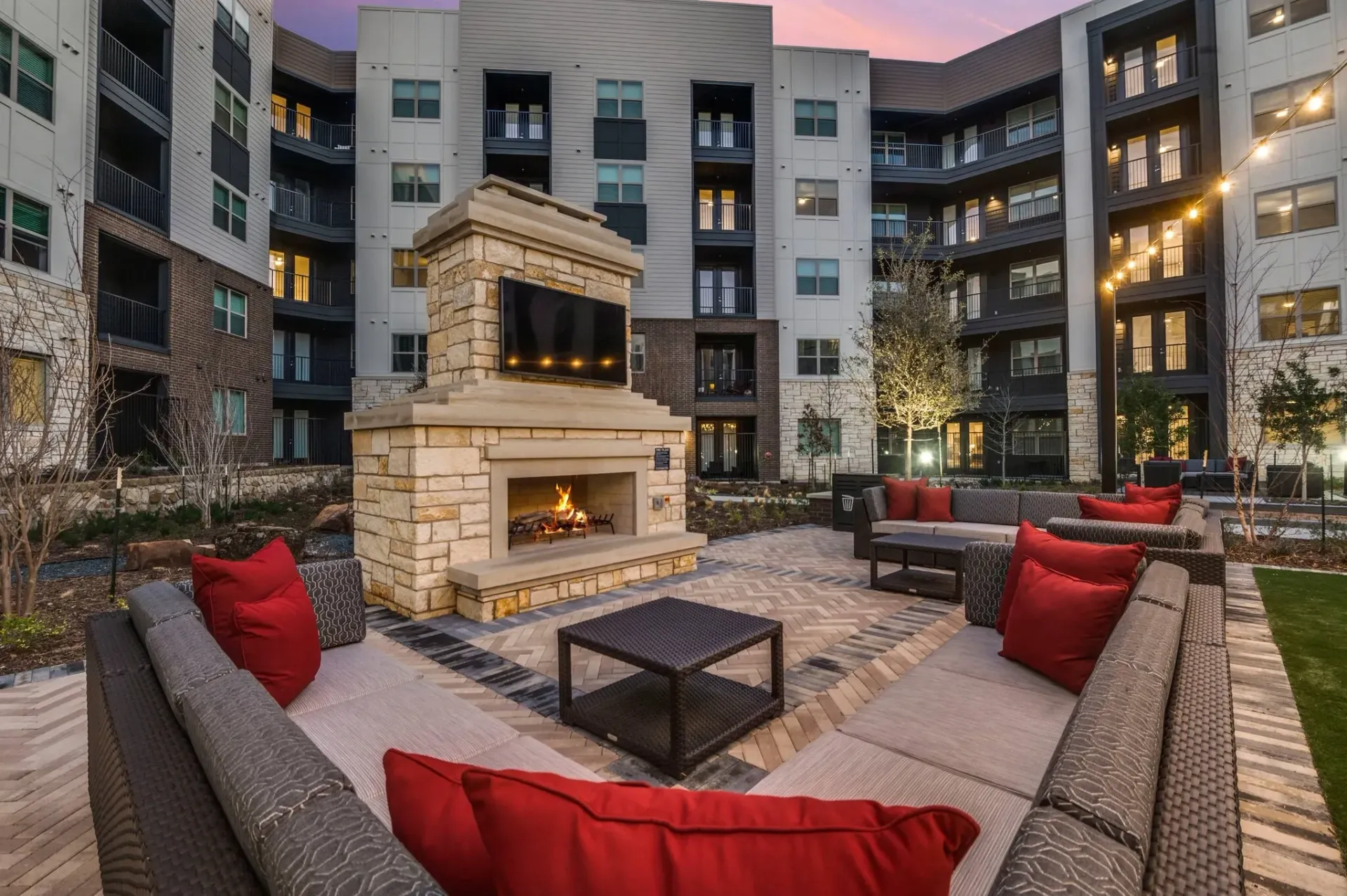 Outdoor courtyard at dusk featuring a stone fireplace with a built-in TV, surrounded by patio furniture with red pillows.
